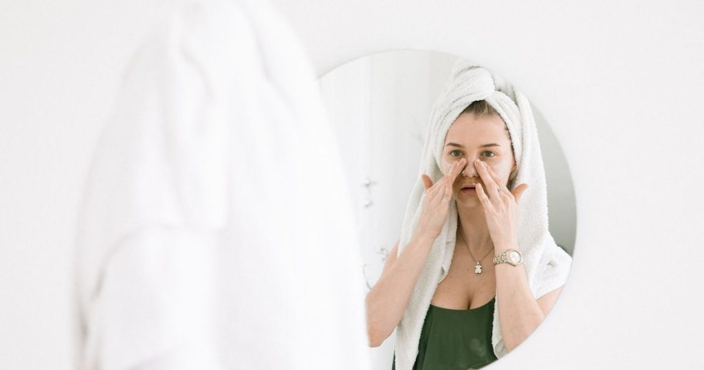 Woman in a towel following a skincare routine at a bathroom mirror